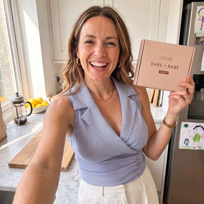 Smiling woman in a blue striped sleeveless wrap top taking a selfie in her kitchen, holding a box of Extra-Boost Sticky Push Up Inserts, a customer review photo