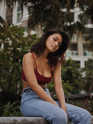 Woman in a burgundy scoop-neck crop top and high-waisted jeans sitting outdoors under palm trees, for a guide to the best nipple cover options for hiding your nipples without wearing a bra