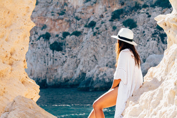 Woman in a white linen cover-up and white sun hat sitting on pale rocks above a turquoise sea, for summer vacation outfit trends to wear braless with nipple covers