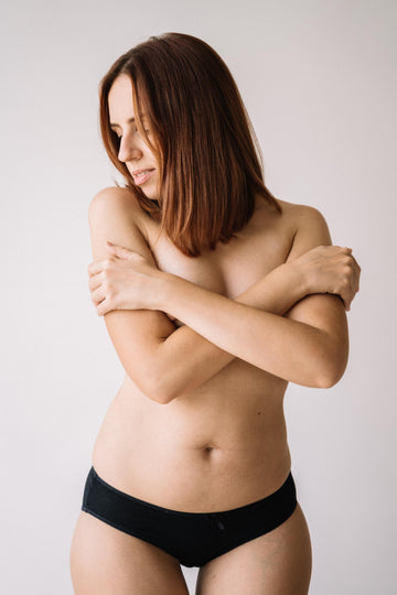 Woman in black briefs with her arms crossed over her bare chest against a white wall, for a guide to whether silicone nipple covers are safe to wear and how to use them
