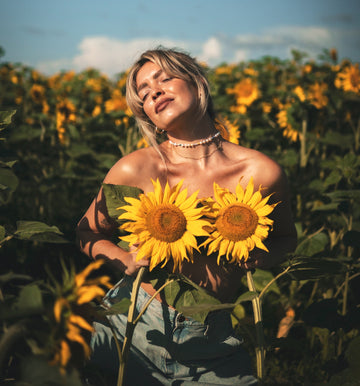 Woman in a sunflower field holding two sunflowers over her chest, for a guide to sustainable, eco-friendly reusable nipple covers you can wash and wear again