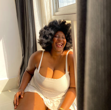 Laughing woman in a white square-neck sundress sitting by a sunny window, for a guide to the best nipple covers for big breasts and larger cup sizes