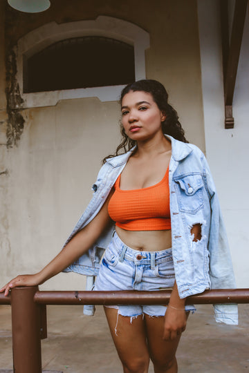 Woman in an orange crop top, faded denim jacket and cut-off shorts leaning on a railing, for tips on reusing nipple pasties and making silicone nipple covers last longer