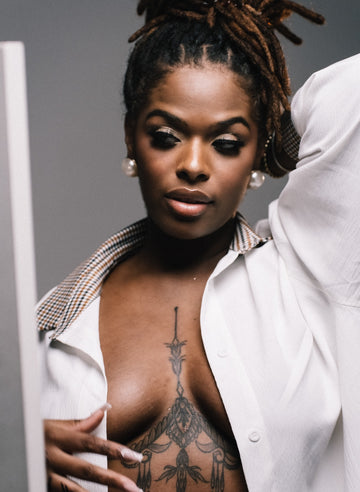 Woman in an open white shirt with a chest tattoo and pearl earrings, one arm raised, for an article on whether nipple covers work and the benefits of silicone nipple covers
