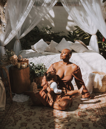 Mother in a rust-colored off-the-shoulder dress breastfeeding her baby in a sunlit bedroom, for an article on nipple covers for breastfeeding moms, comfort and confidence