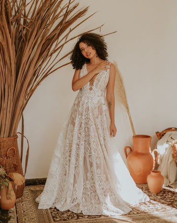 Woman with curly hair in a white lace gown standing in a boho room with pampas grass, for a guide to summer wedding dresses and wearing nipple covers under them