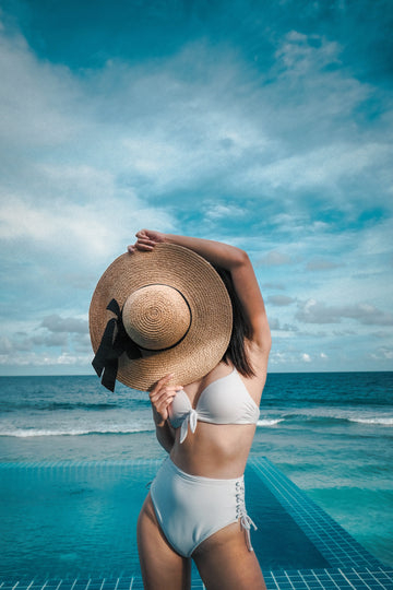 Woman in a white bikini holding a wide straw sun hat over her face beside an infinity pool and the ocean, for a guide to waterproof adhesive silicone nipple covers under swimwear