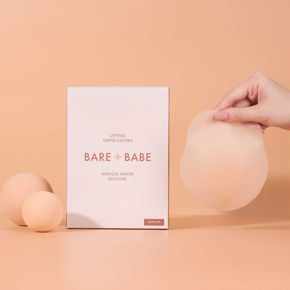 Lifting Nipple Covers (Two-Tabs) box in medical-grade silicone, labelled With Lift, beside a hand holding up one two-tab reusable silicone nipple cover on a peach background