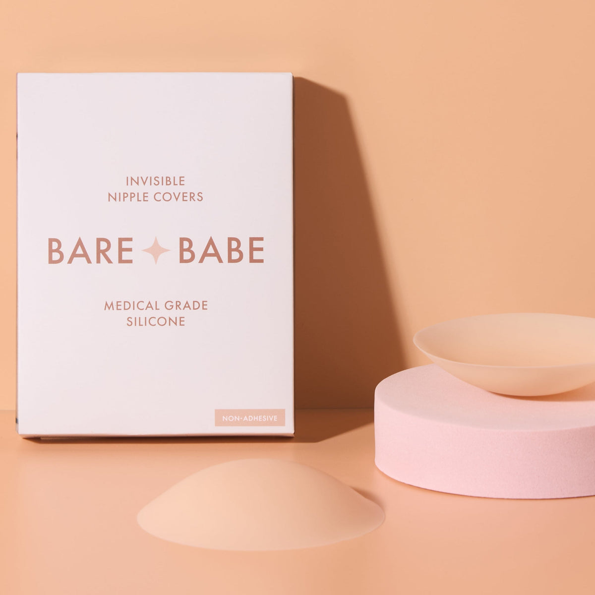 Non-Adhesive Nipple Covers in Creme: two round, non-sticky reusable silicone nipple covers, one on a pink stand and one dome-side up, beside the Bare Babe medical-grade silicone box.
