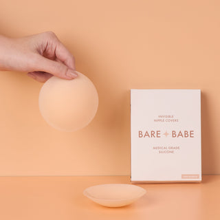 Non-Adhesive Nipple Covers in Creme: a hand holds up a round, grippy, non-sticky silicone nipple cover, with a second cover and the Bare Babe non-adhesive box on a peach backdrop.