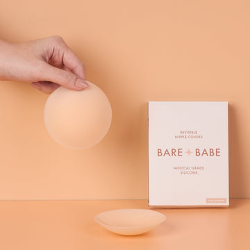 Non-Adhesive Nipple Covers in Creme: a hand holds up a round, grippy, non-sticky silicone nipple cover, with a second cover and the Bare Babe non-adhesive box on a peach backdrop.