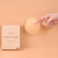 Adhesive Nipple Covers in Honey: a fingertip touches a round sticky silicone nipple cover floating beside the Bare Babe invisible nipple covers box on a soft peach backdrop.