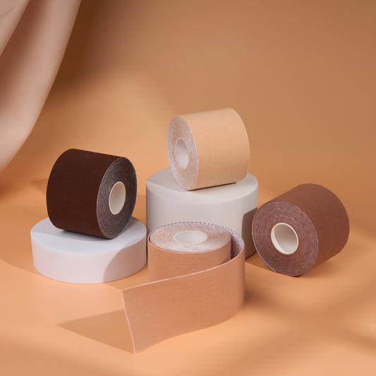 Boob Tape Kit rolls in four shades stacked on white display blocks, one roll unspooling to show the soft fabric boob tape that gives real lift and support under backless and strapless outfits.