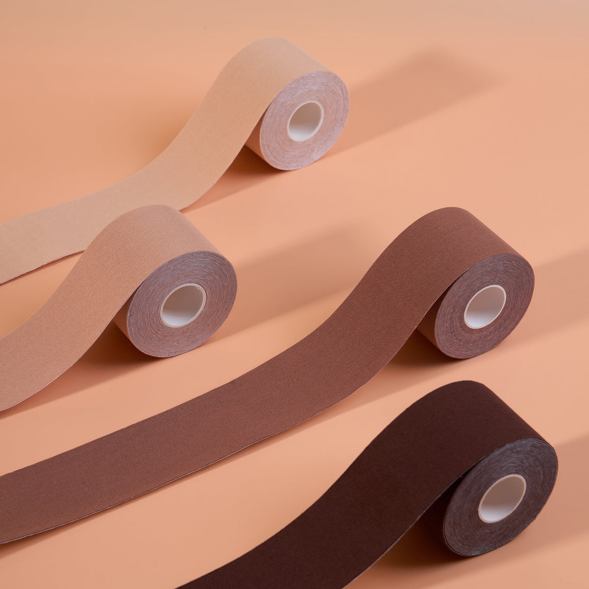 Boob Tape Kit rolls in four shades unrolled side by side, showing the matte fabric finish of the breast lift tape worn for lift and support under strapless, backless and plunge necklines.