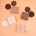 Close-up of Satin Nipple Pasties boxes labeled disposable satin nipple covers, 20 pairs, in four shades, with round and flower nipple pasties spread around them.