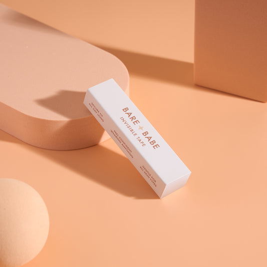 Invisible Lingerie Tape box resting on peach display props, a slim pack of pre-cut double-sided clothing tape strips marked secure fit all day long and gentle for all skin types.