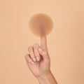 Adhesive Nipple Covers: a single round silicone nipple cover balanced on a fingertip, showing how thin and matte the reusable nipple pasty is, against a peach background.