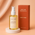 Boob Tape Remover Oil: a 100 ml / 3.4 fl oz spray bottle of golden oil on a pedestal beside its rust-red Body Tape Remover Oil box, for gentle removal of boob tape, body tape and lingerie tape adhesive.