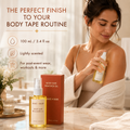 Boob Tape Remover Oil in use: a woman in a cream robe sprays the oil onto her shoulder at a marble vanity. 100 ml / 3.4 fl oz, lightly scented, for taking off body tape after events, workouts and more, on intact skin.